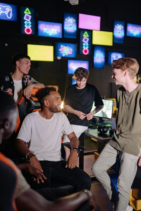 Five young men in a game room, talking and laughing. Decorated with neon signs and screens.