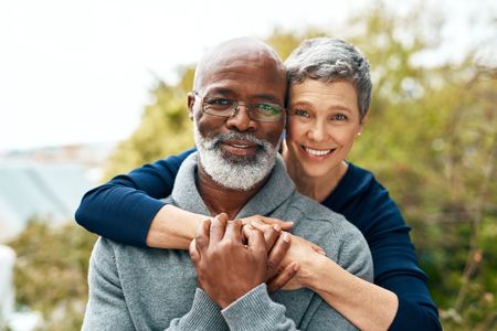 Smiling couple embracing outdoors. Woman's arms around man, hands clasped. Both have gray hair.