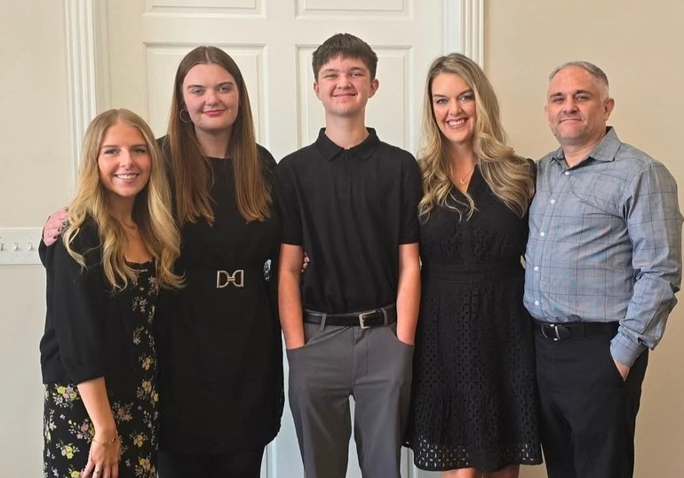 Family of four at a table, seaside. Man, woman, teen boy, and girl smile. Red dress, blue table settings.
