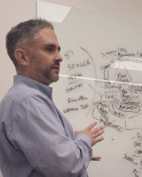 A man presenting to a group seated around a conference table in a room with a TV screen.