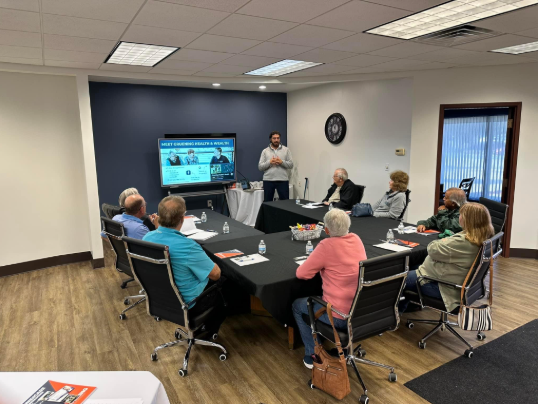 A man presenting to a group seated around a conference table in a room with a TV screen.