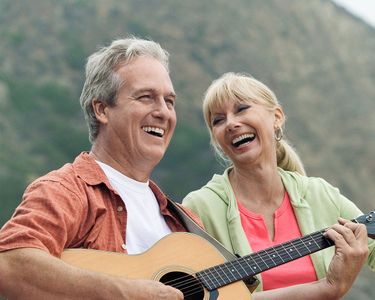 Man playing guitar, smiling; woman laughing nearby. Outdoors, sunny day.