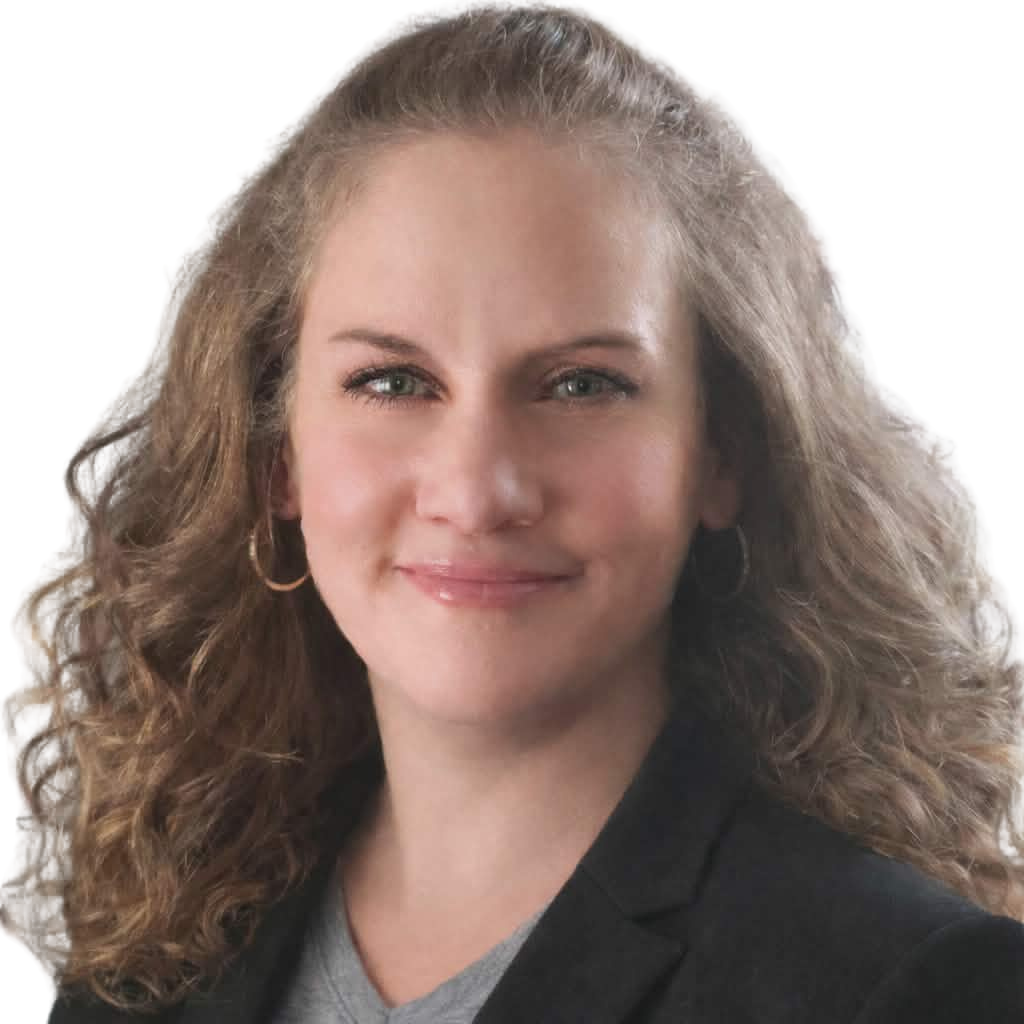 Woman with curly brown hair, wearing a grey shirt and black blazer, smiling at camera.