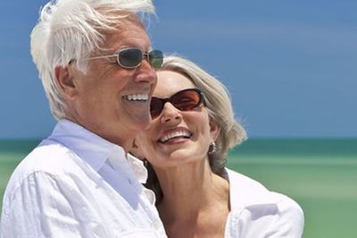 Couple in sunglasses smiling by the ocean. White shirts, blue sky.