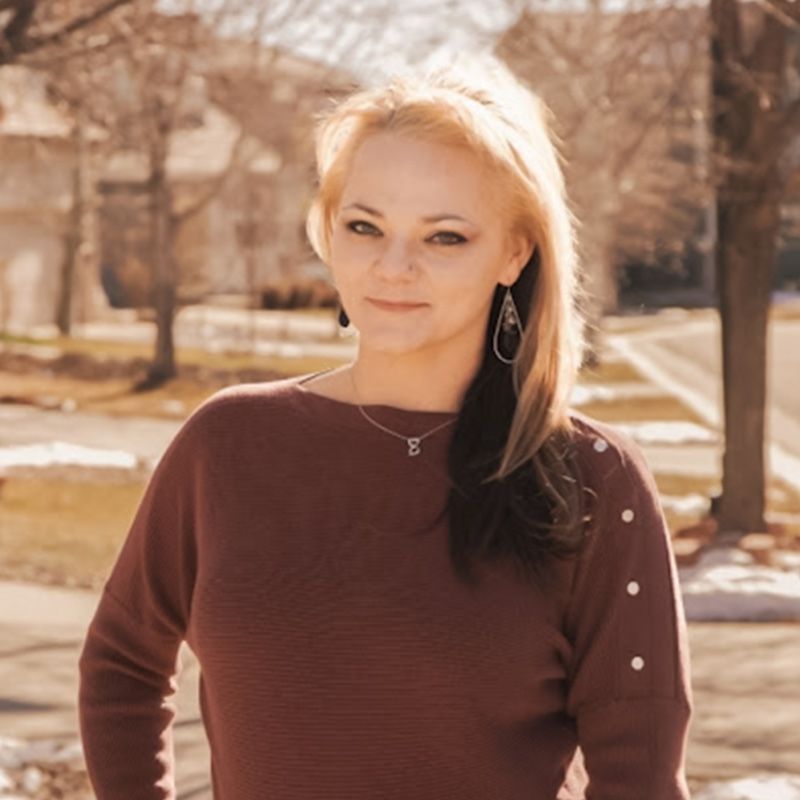Woman with blonde hair, maroon sweater, standing outside with a neighborhood background.