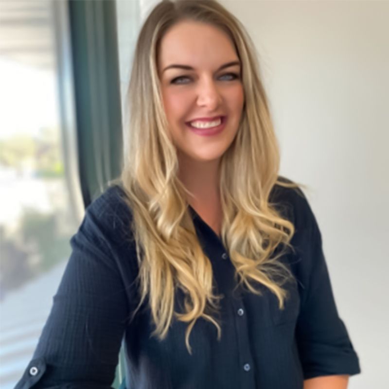 Woman with long blonde hair smiles, wearing a black button-down shirt, near a window.