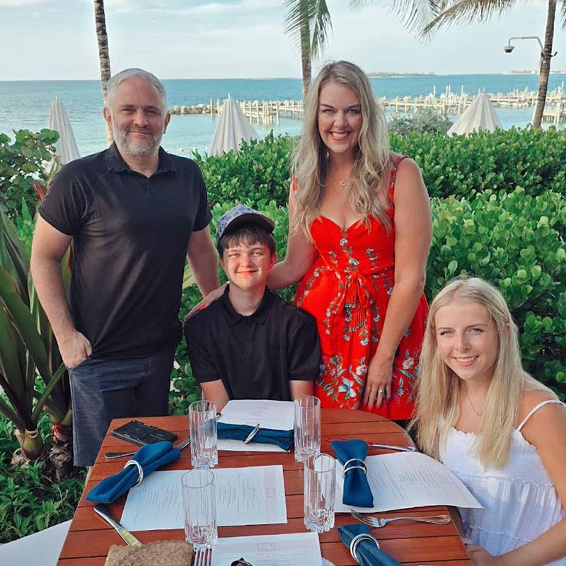 Family of four at a table, seaside. Man, woman, teen boy, and girl smile. Red dress, blue table settings.