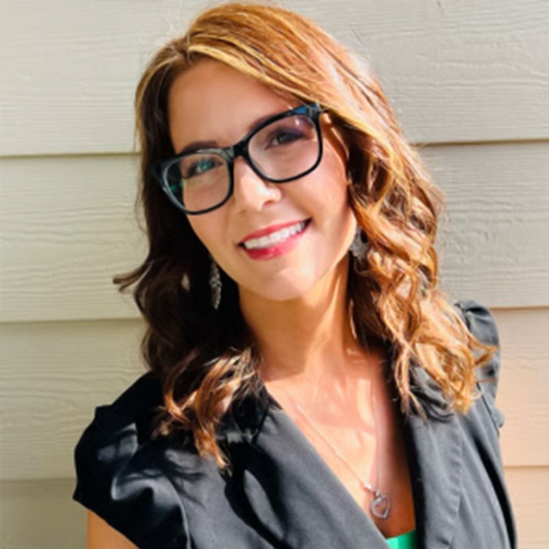 Woman with glasses smiles, standing in front of a light-colored building, wearing a black blazer.