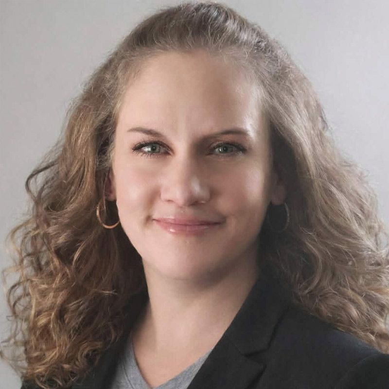 Woman with curly brown hair, wearing a grey shirt and black blazer, smiling at camera.