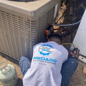 HVAC technician working on an air conditioning unit. Gray unit, white shirt with logo, crouching outdoors.