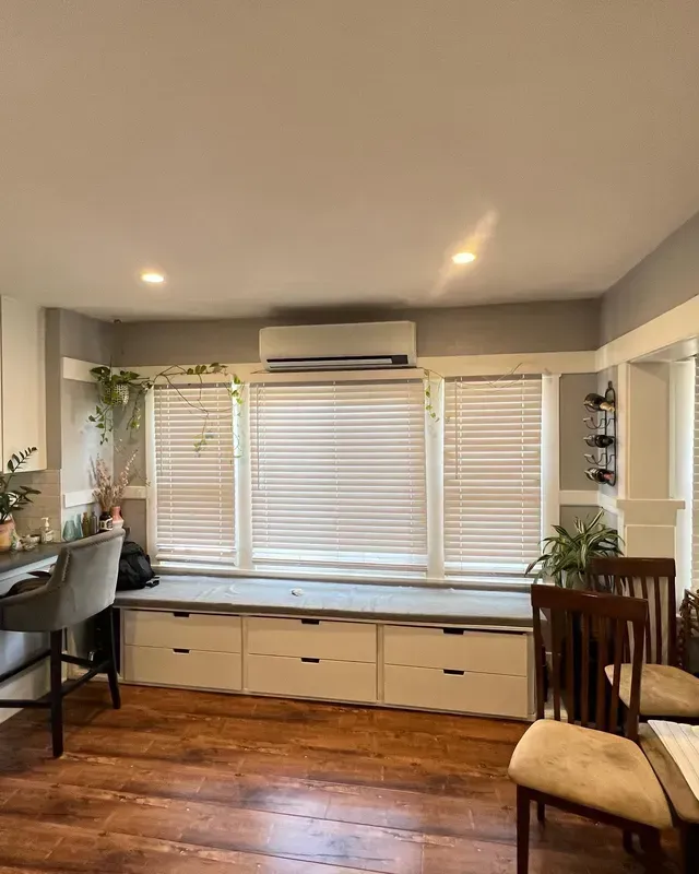 A kitchen with a window seat, blinds, and drawers below. Wood floors, grey walls, and hanging plants.