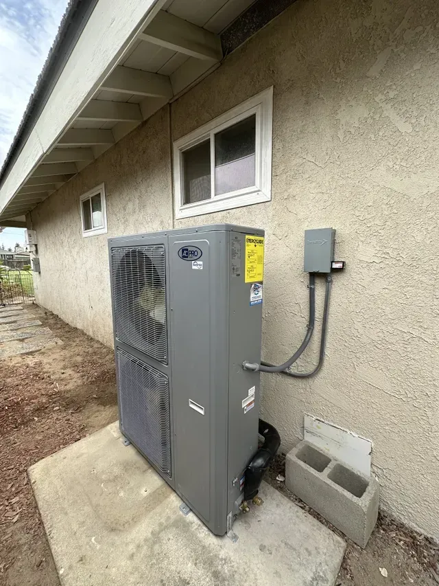 Exterior view of a gray HVAC unit beside a stucco building with a window and electrical box.