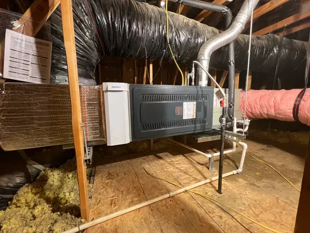 HVAC unit in attic, black and silver, connected to ducts. Insulation visible. Wooden beams in background.