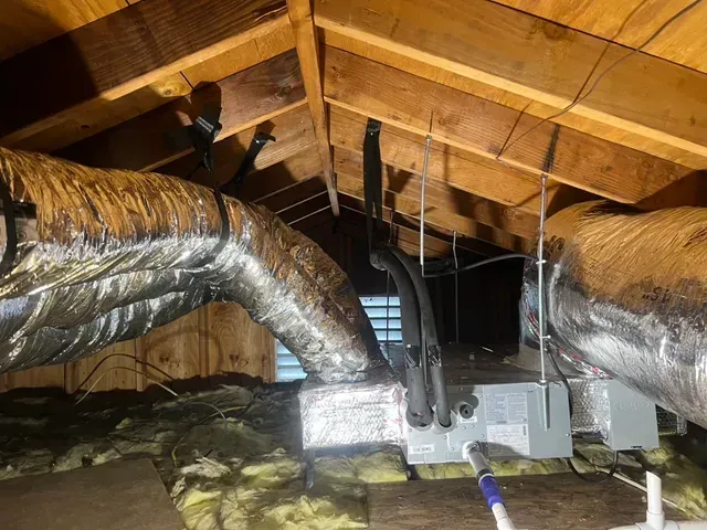 HVAC ductwork and unit in an attic with insulation and wooden beams.