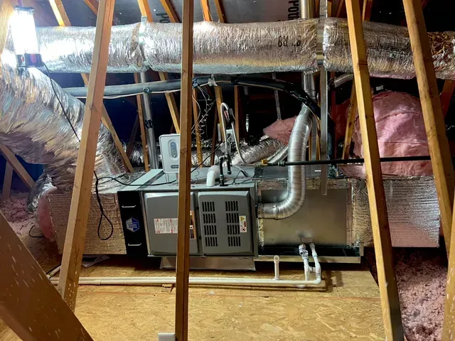 HVAC unit in an attic with ductwork and insulation, surrounded by wooden framing.