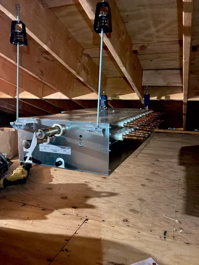 HVAC unit suspended in attic, supported by metal rods attached to wooden beams.