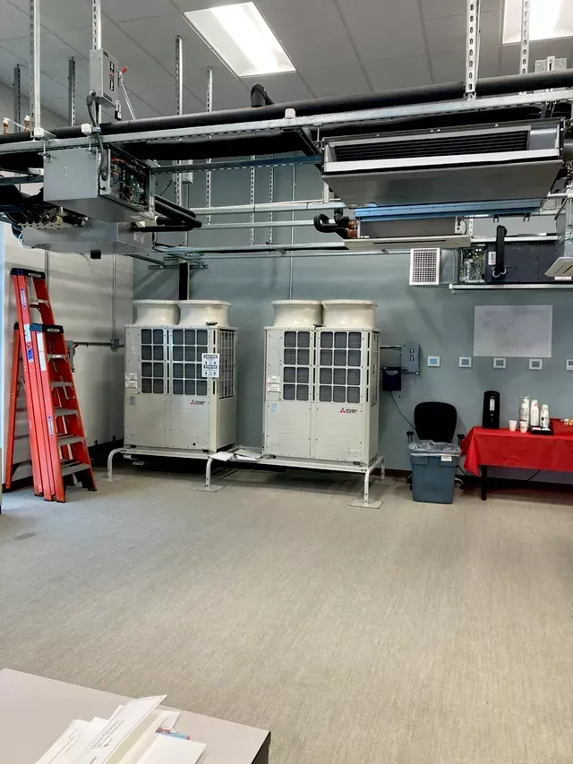 Two industrial cooling units in a room, with a ladder and overhead pipes.
