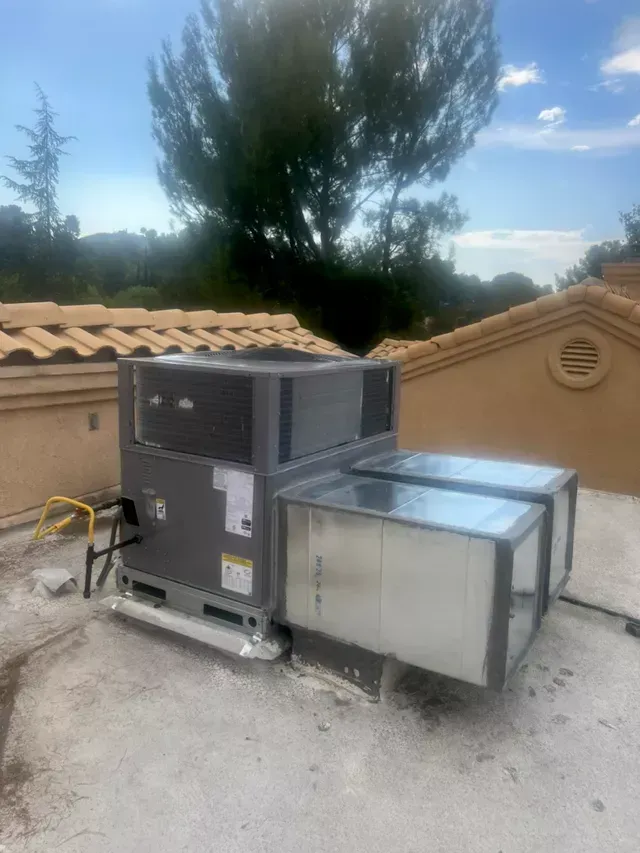 Rooftop air conditioning unit with ductwork on a tile roof, blue sky backdrop.