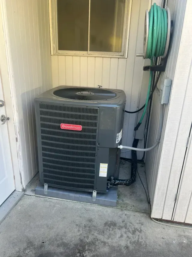 Outdoor AC unit near a window with a green hose, set against a white wall.