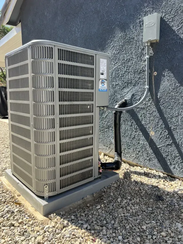 Gray HVAC unit next to a gray wall and a gravelly ground.