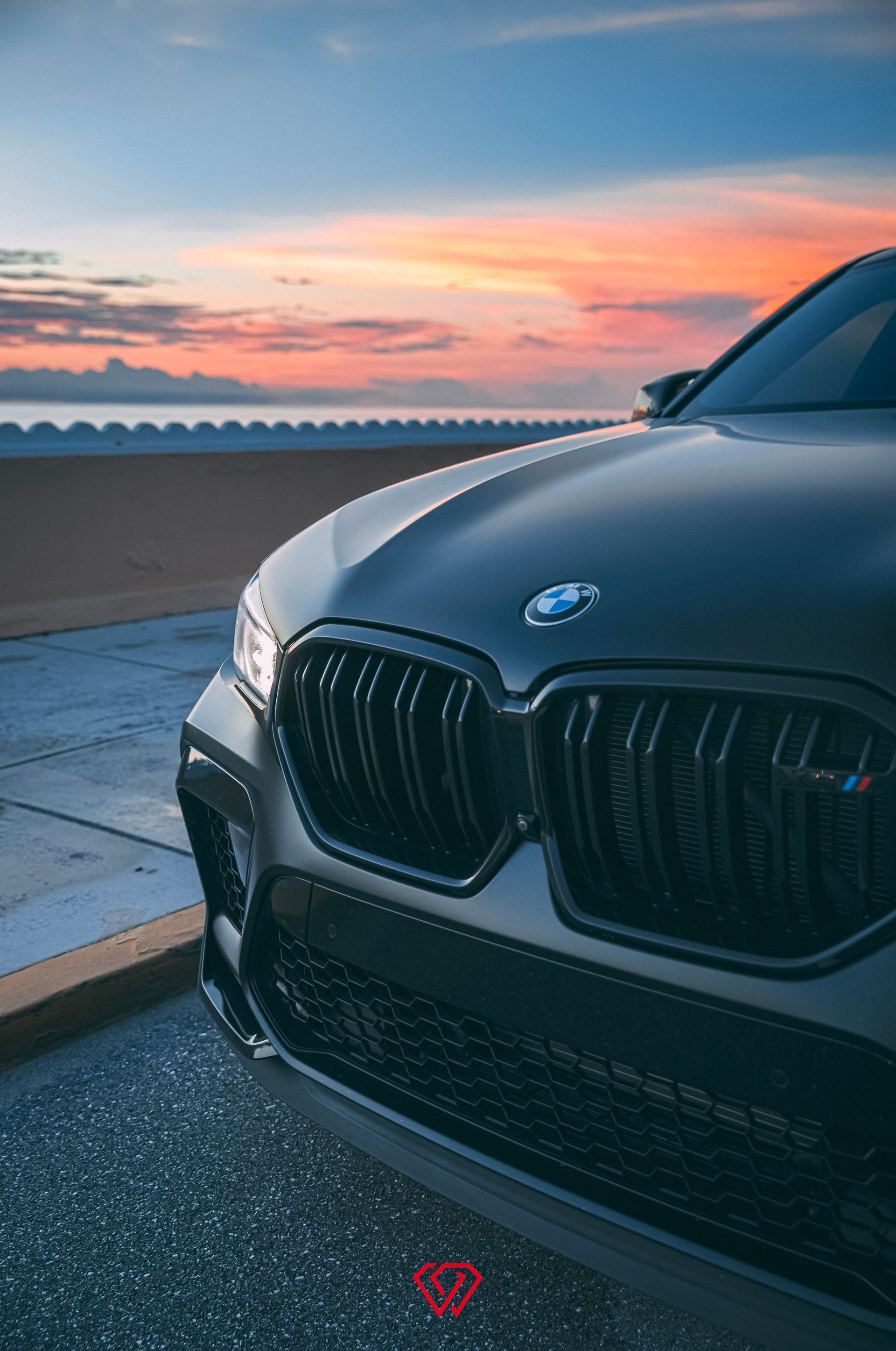 Dark gray BMW SUV, front view, at sunset near the water. Black grille and logo are visible.