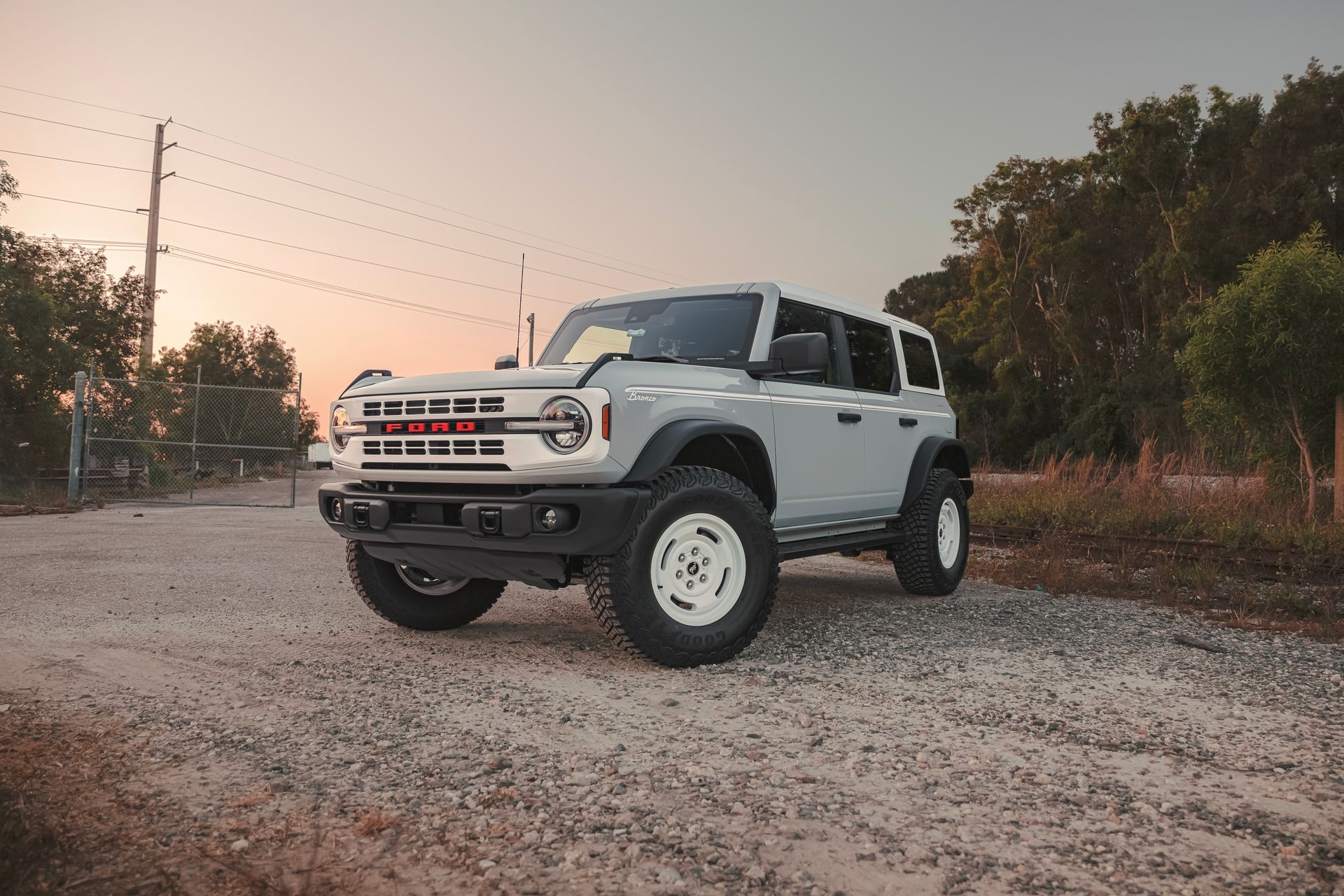 Light gray Ford Bronco parked on a gravel road with white wheels, sunset background.
