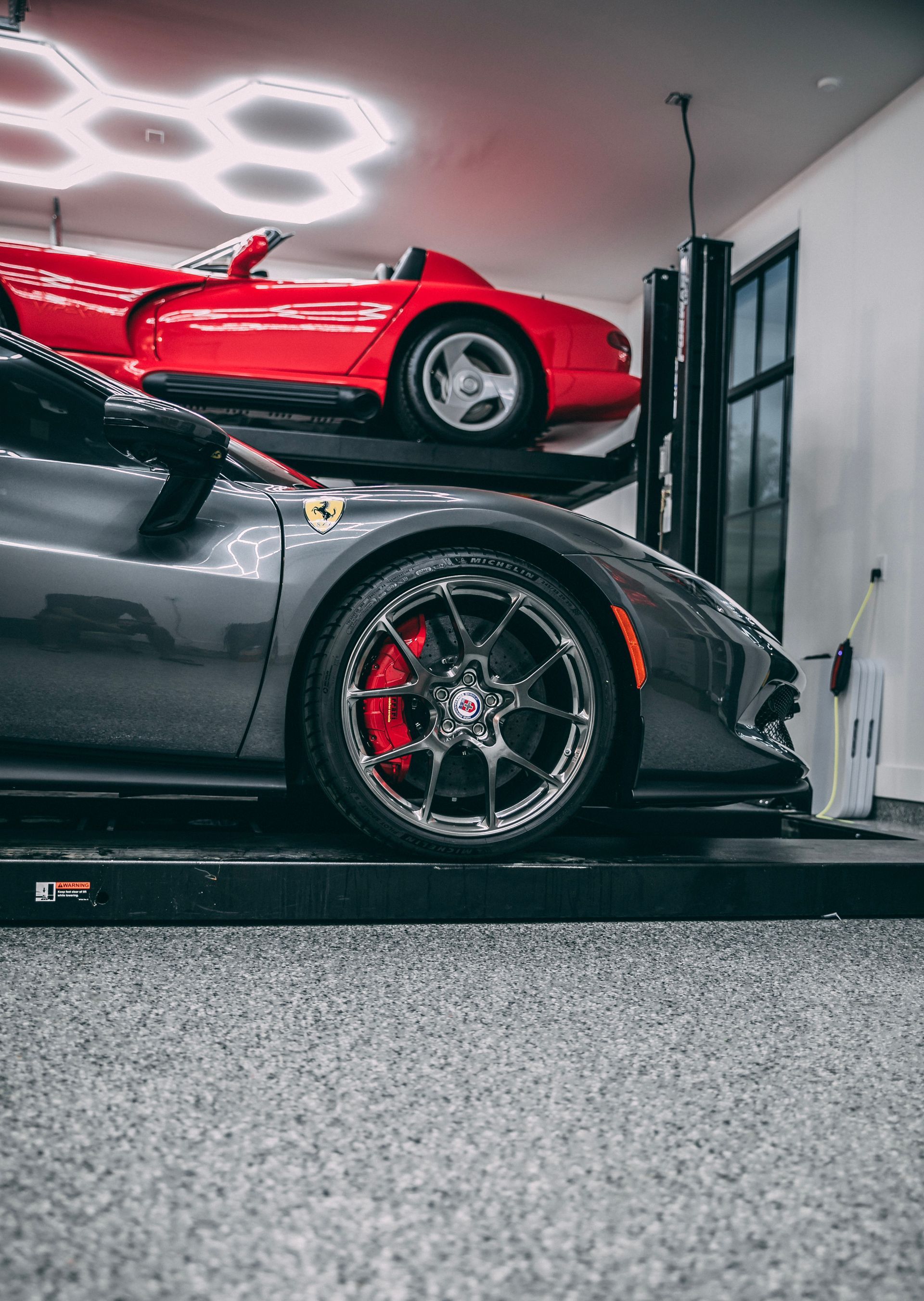 Two sports cars, a grey Ferrari and red Dodge Viper, on a car lift in a garage with epoxy flooring.