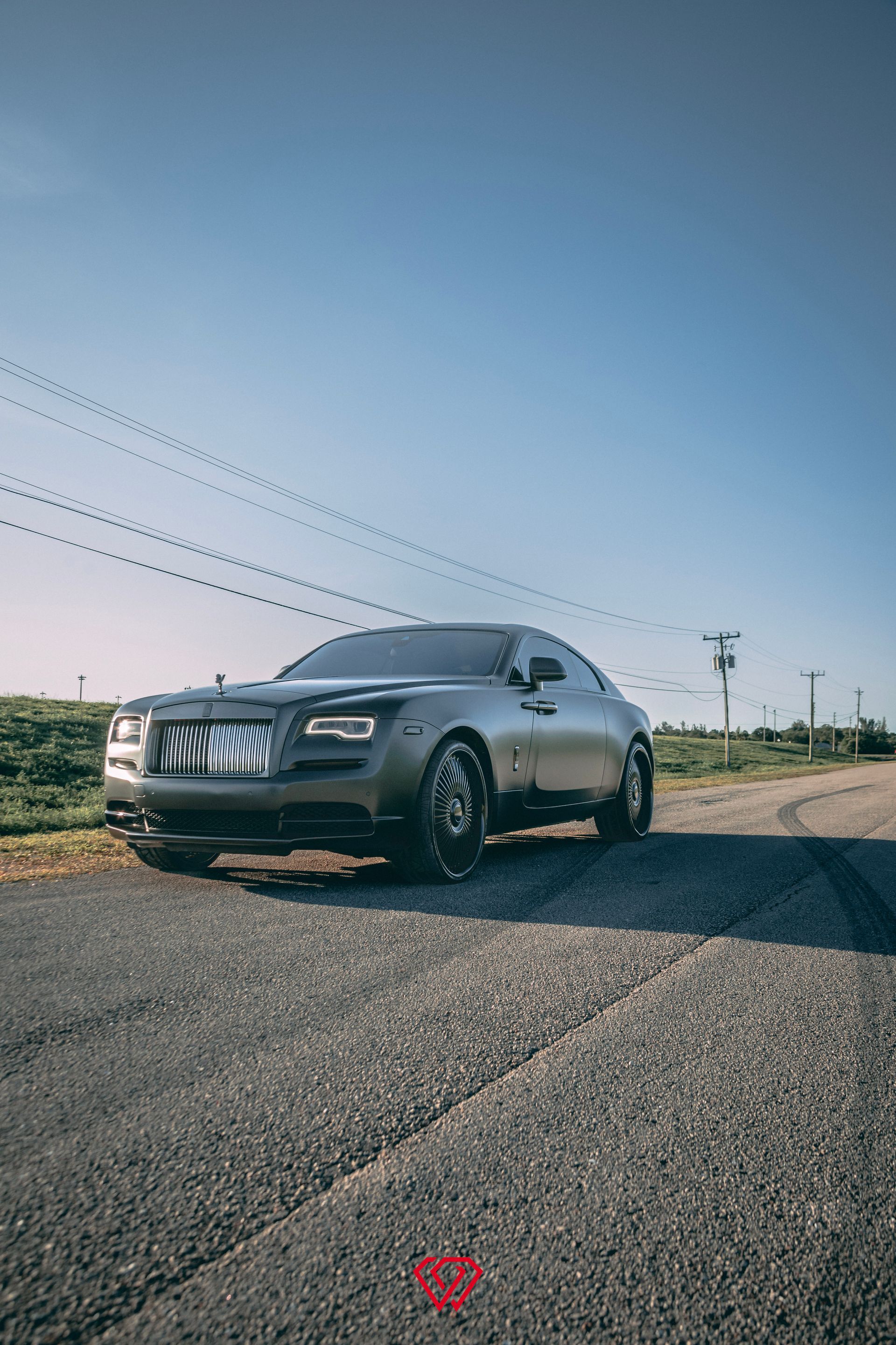 Gray Rolls Royce coupe parked on a gravel road under a blue sky, power lines overhead.