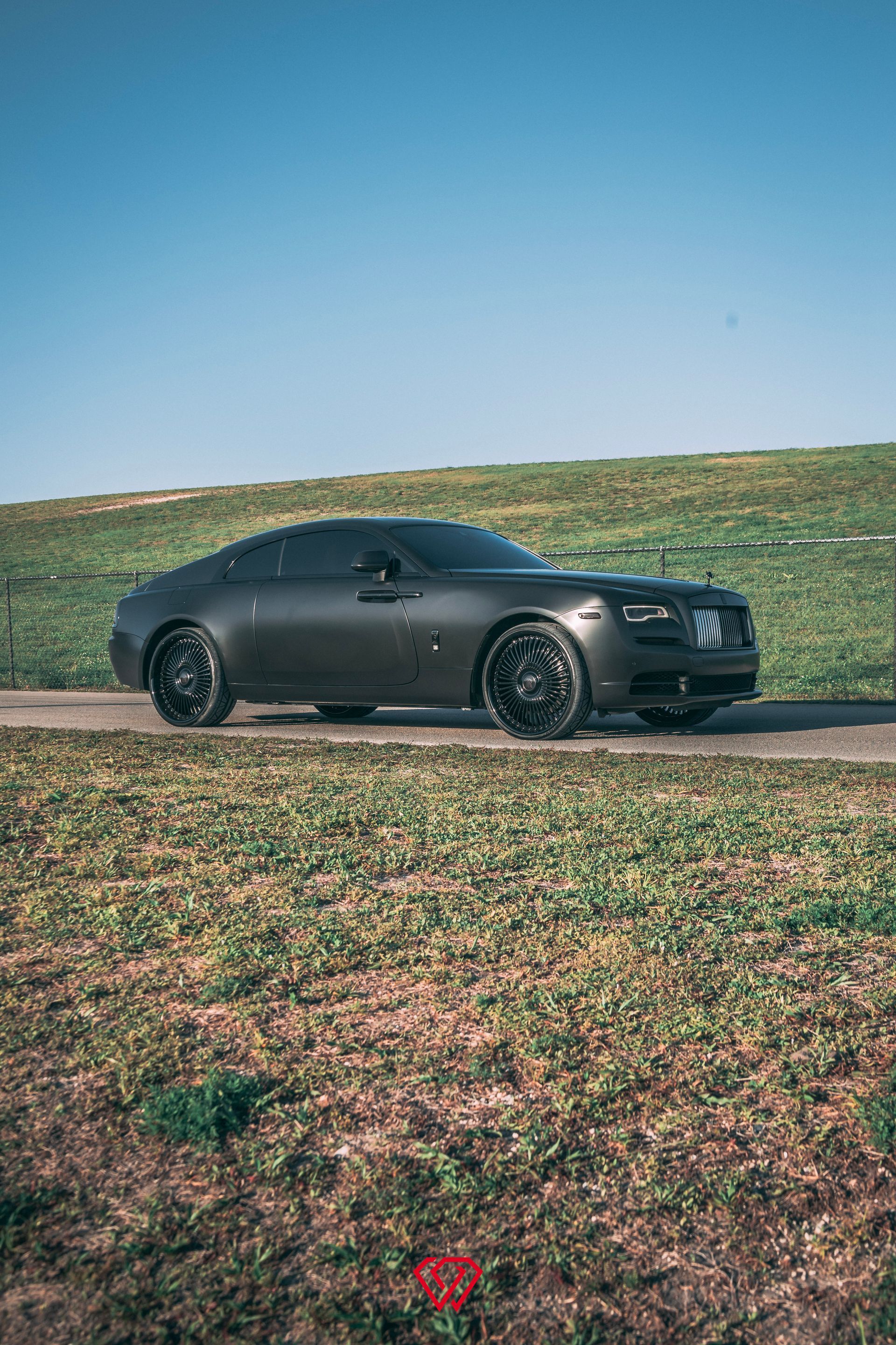 Dark gray Rolls-Royce coupe parked on a road beside a grassy hill under a blue sky.