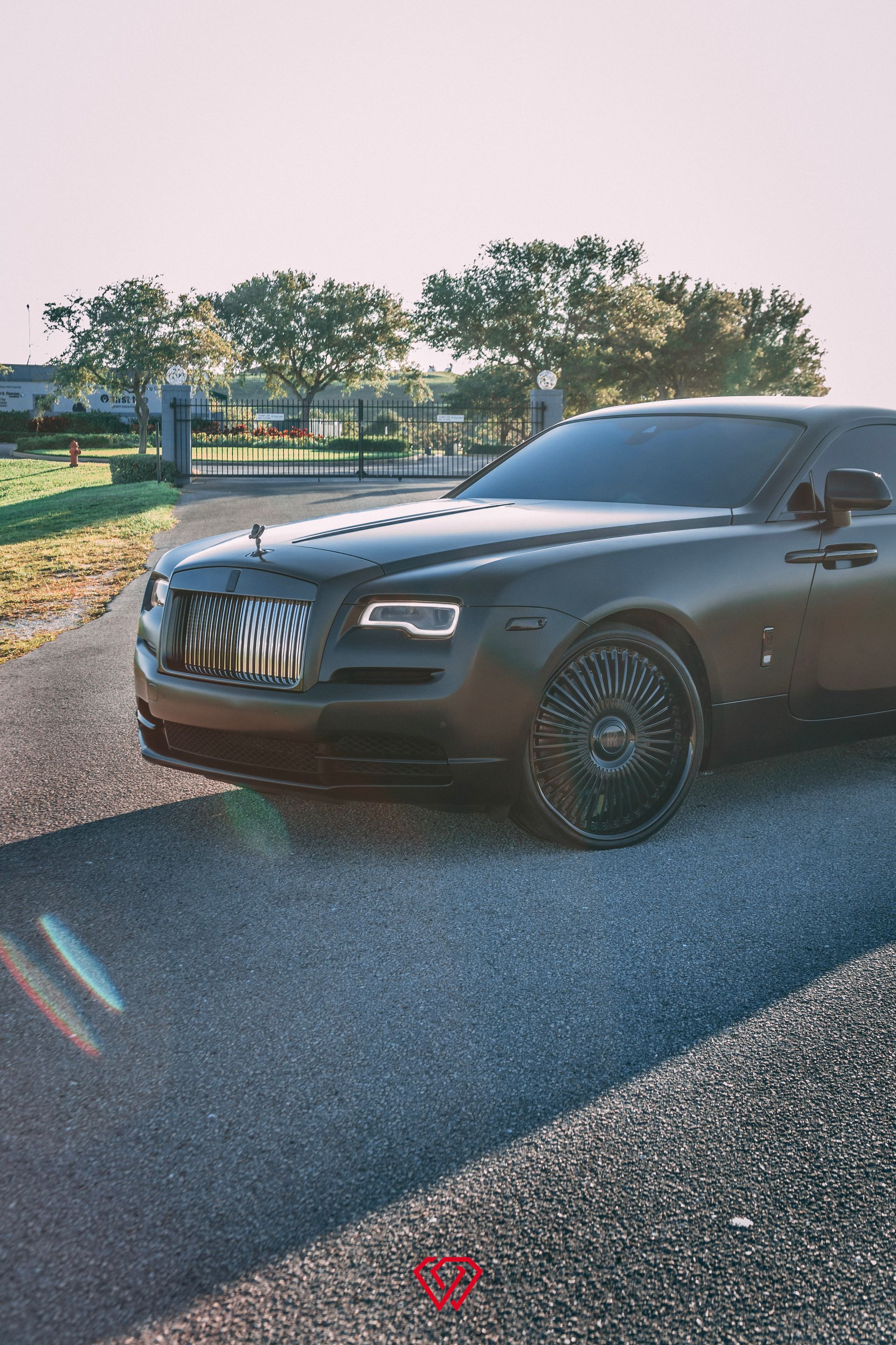 Black Rolls-Royce parked on a driveway with a gate and trees in the background.