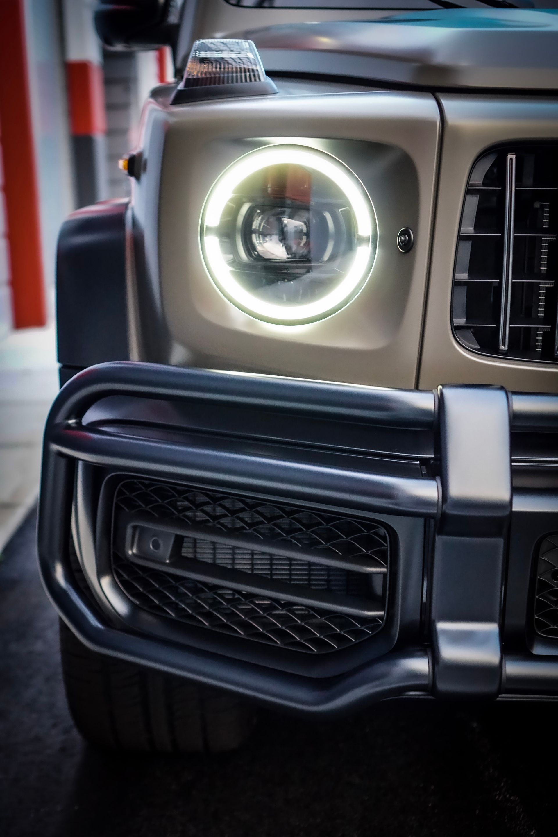 Close-up of a tan Mercedes-Benz G-Class front. Headlight and black grill are prominent.