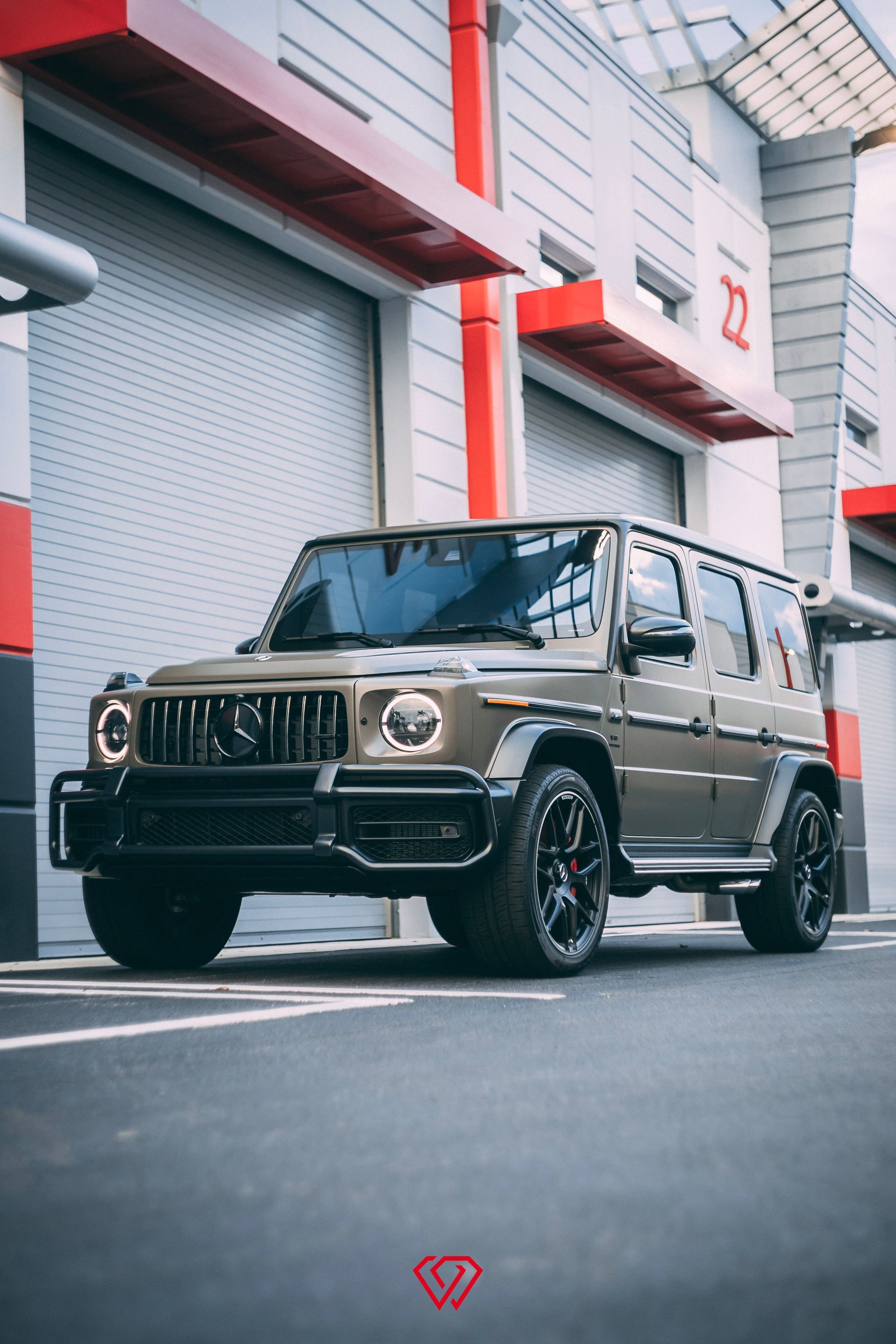 Olive-green Mercedes-Benz G-Class SUV parked in front of a modern building with gray garage doors and red accents.