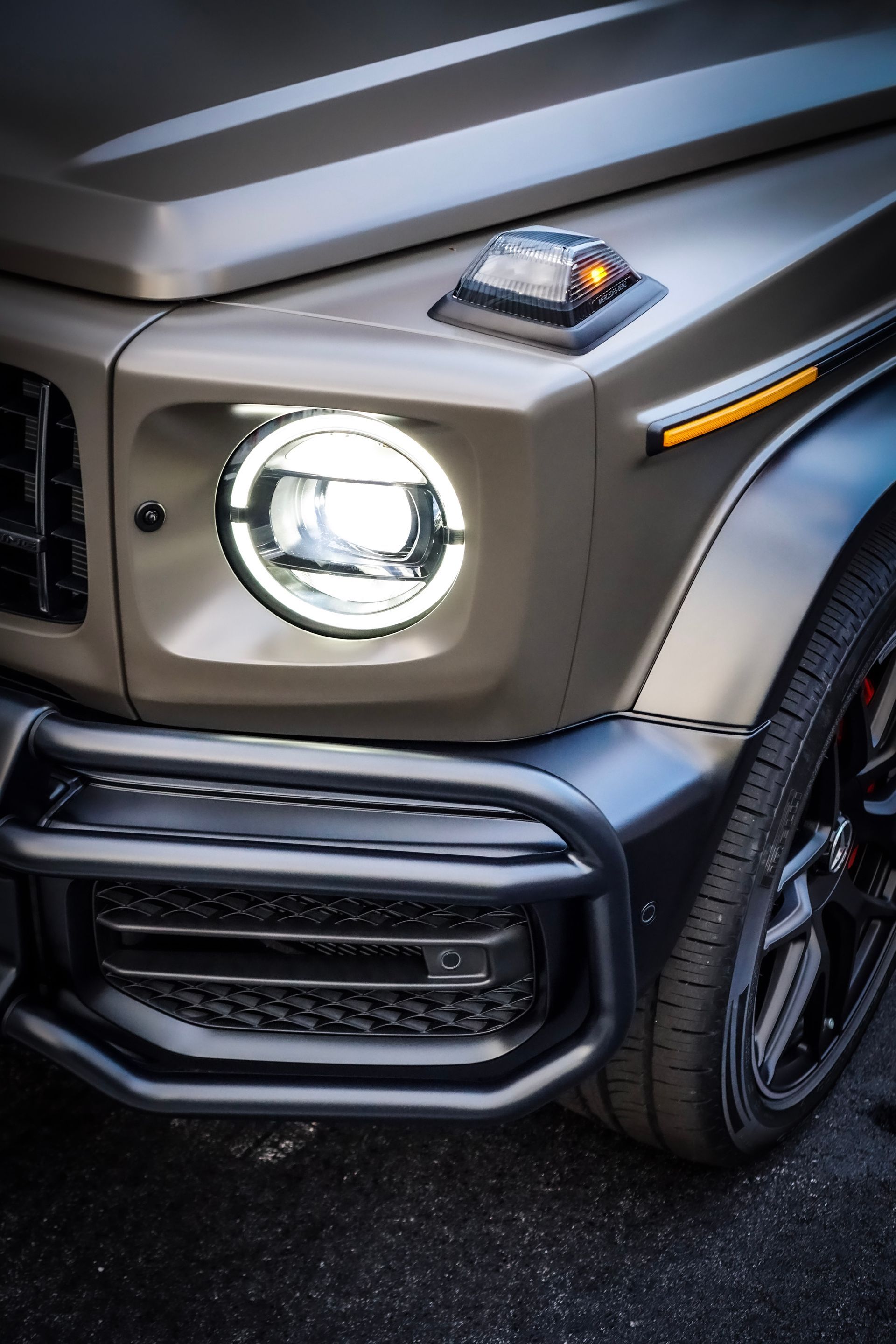 Close-up of a matte gray Mercedes-Benz G-Class SUV's front, highlighting the headlight and bumper.
