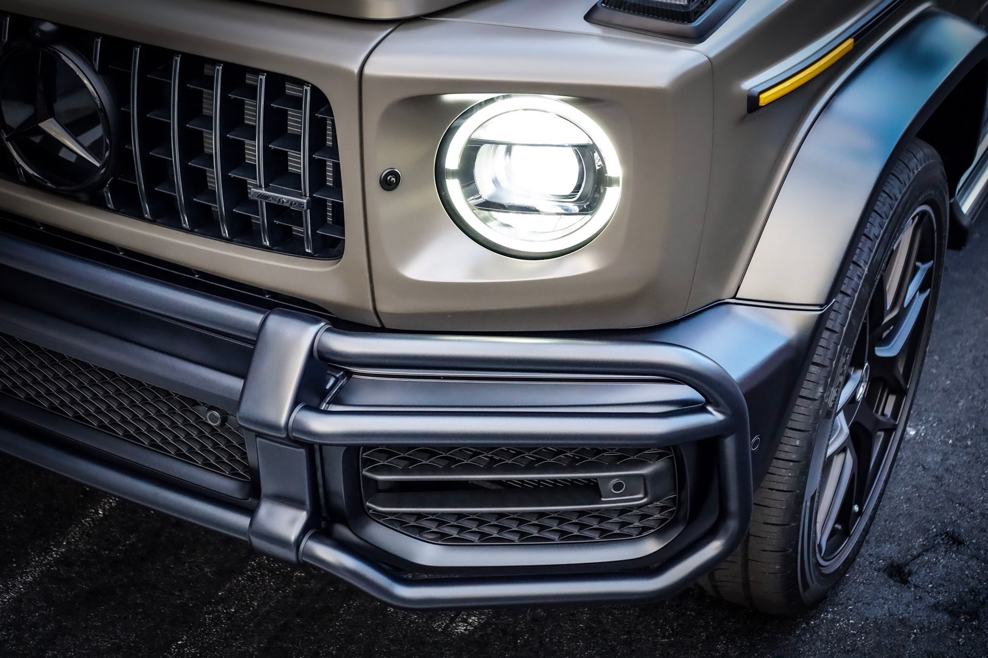 Close-up of a tan Mercedes-Benz G-Class SUV's front end, with a bright headlight and black grille and bumper.