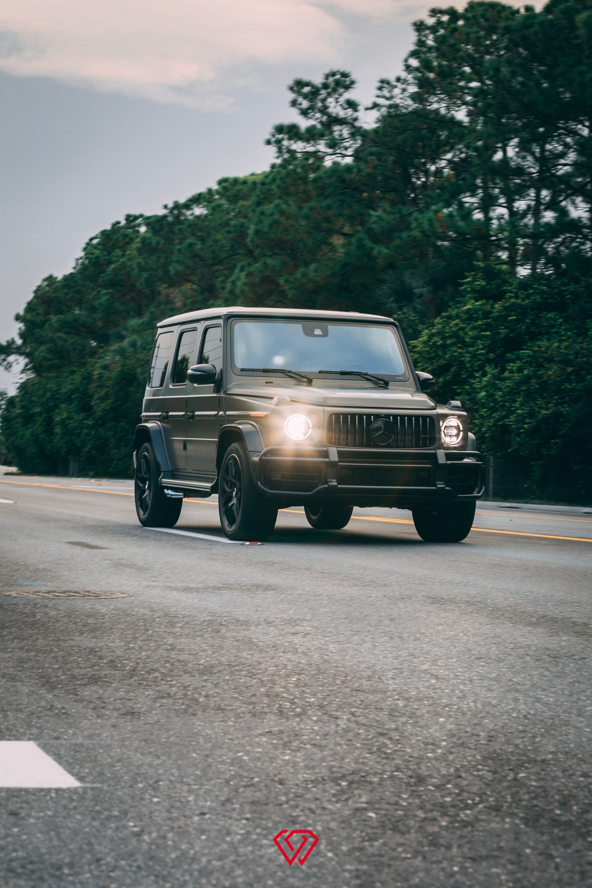 Dark gray Mercedes-Benz G-Class SUV driving on a road with trees in the background.