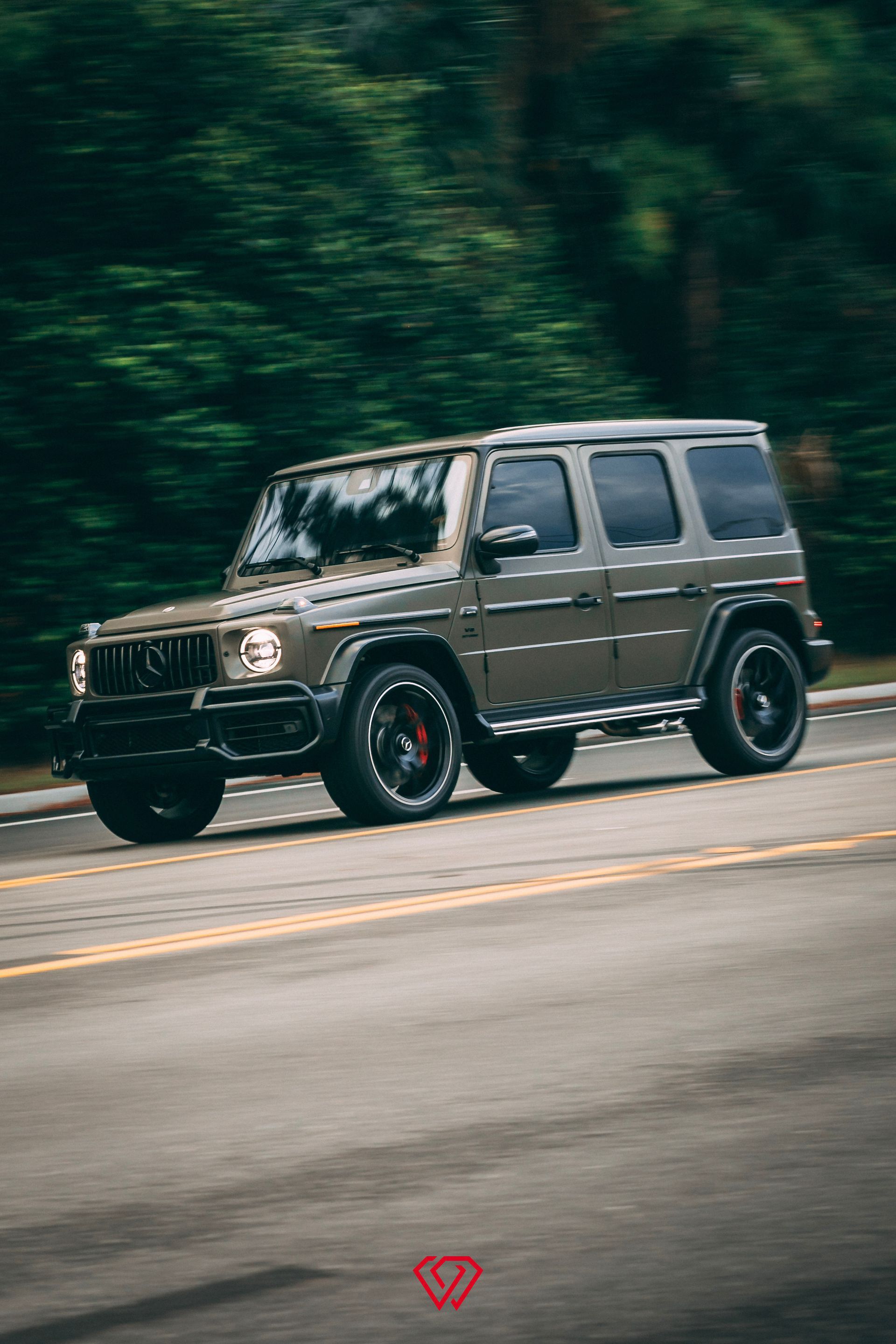Green Mercedes-Benz G-Class SUV driving on a road with blurred background of trees.