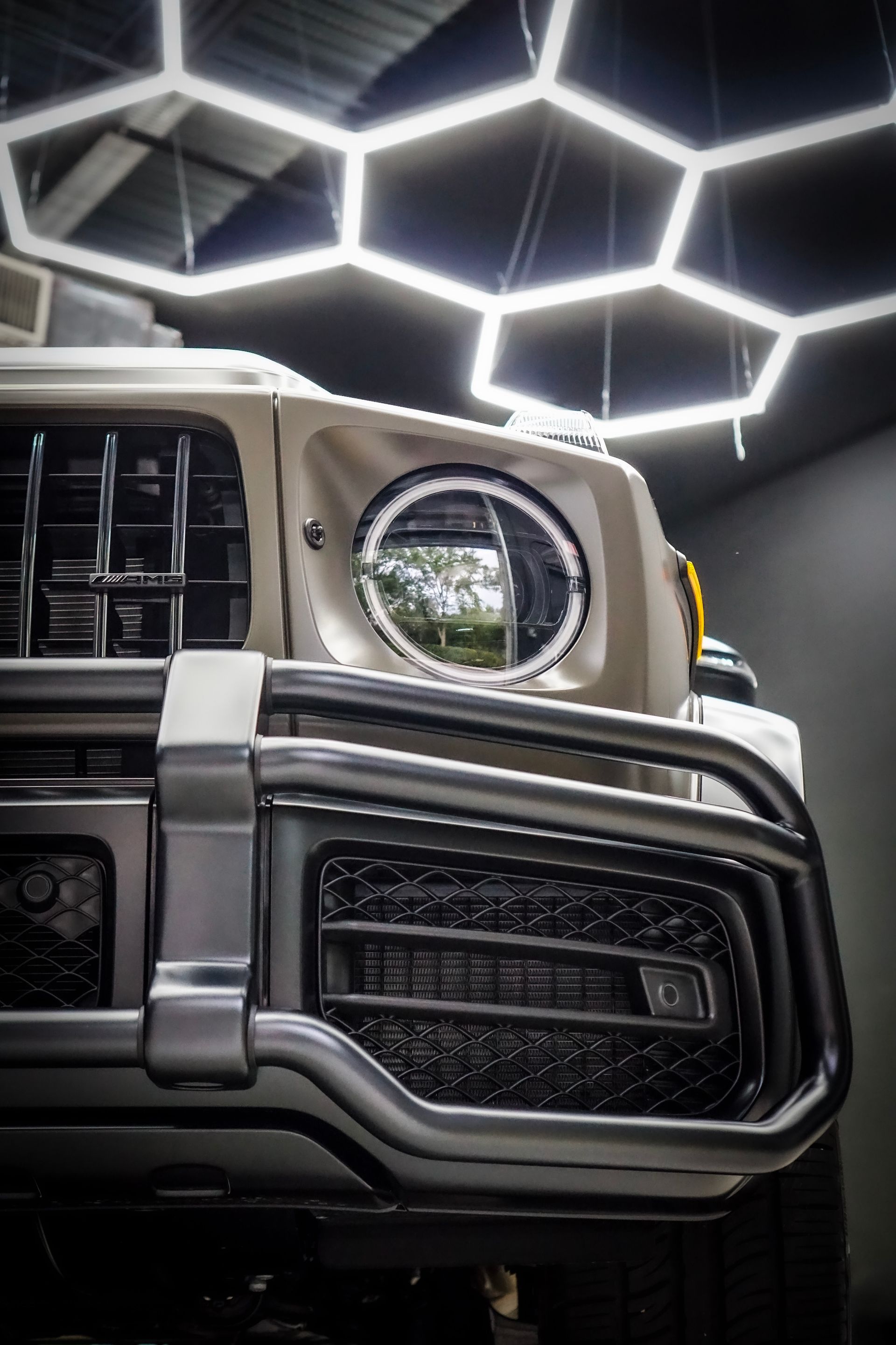 Close-up of a white SUV's front, highlighting its grille, headlights, and a protective bar, with hexagonal lights in the background.