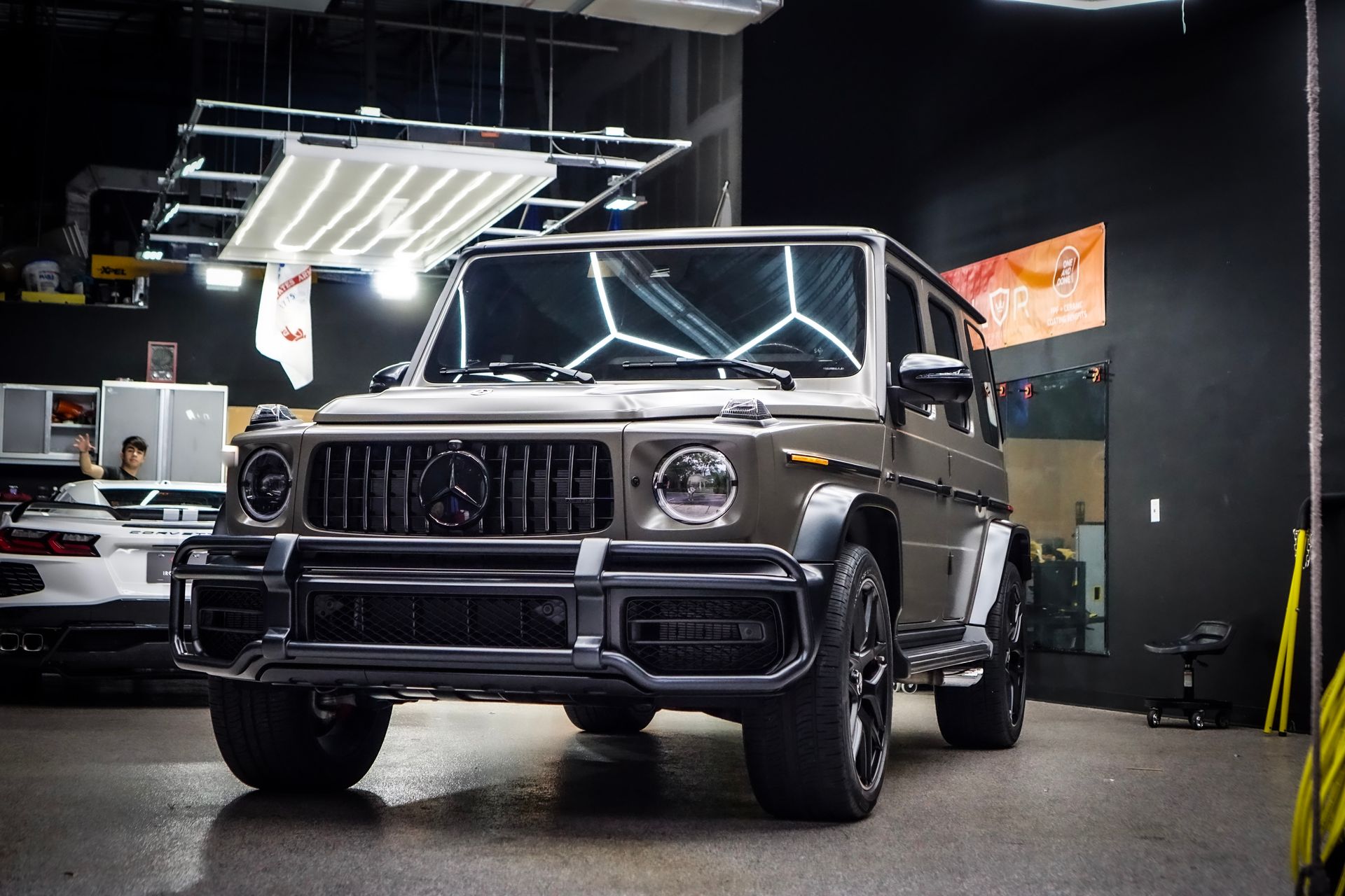 Gray Mercedes-Benz G-Wagon SUV in a well-lit shop with a white sports car visible in the background.
