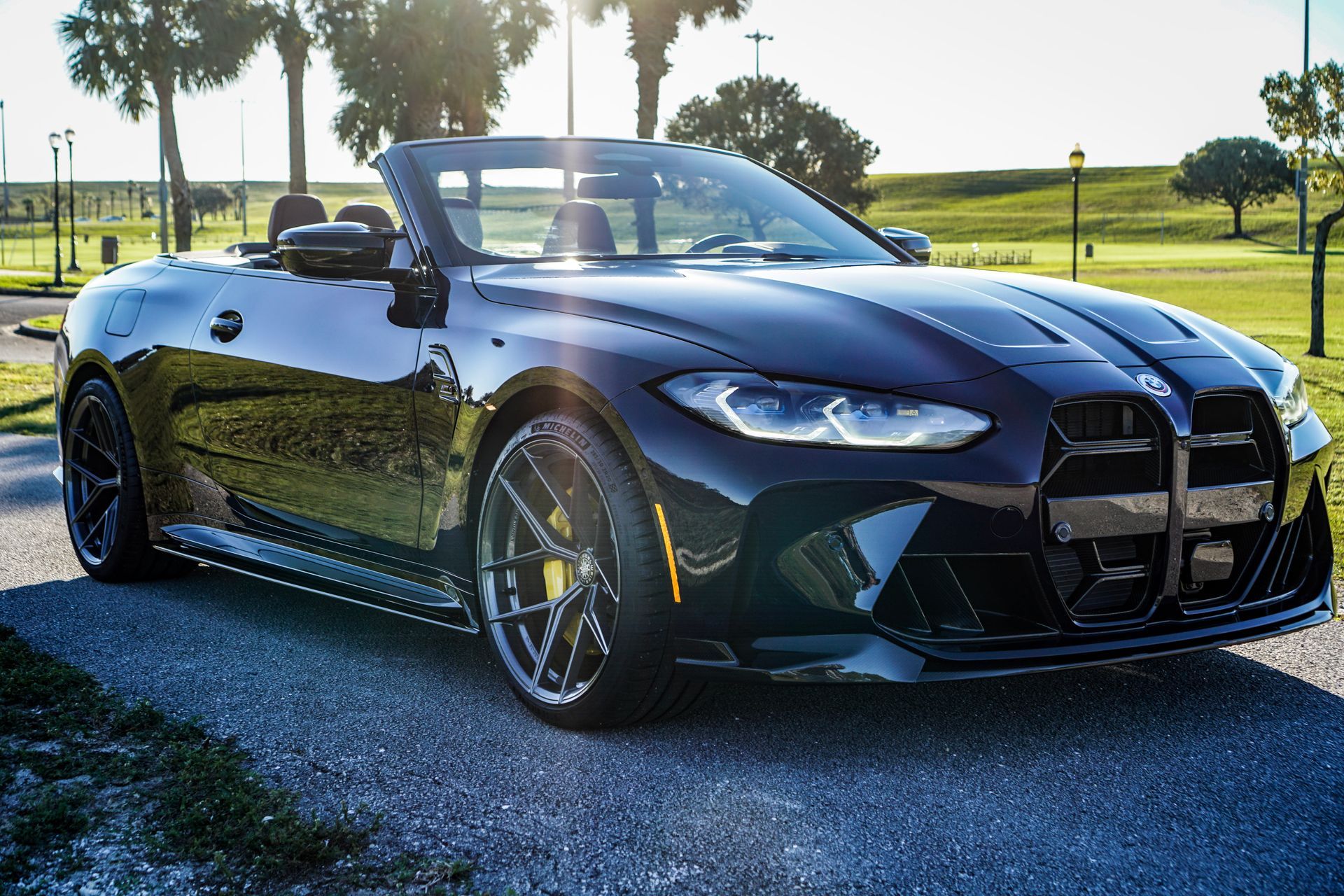 Black BMW M4 convertible parked on a paved road, sunlight in background, golf course setting.