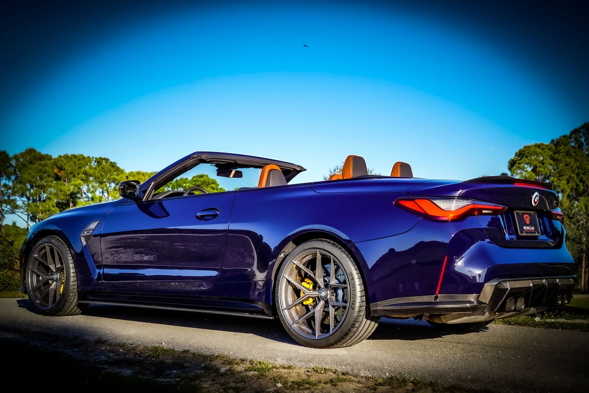 Dark blue BMW M4 convertible with top down, brown interior, parked on a road with trees and a blue sky in the background.