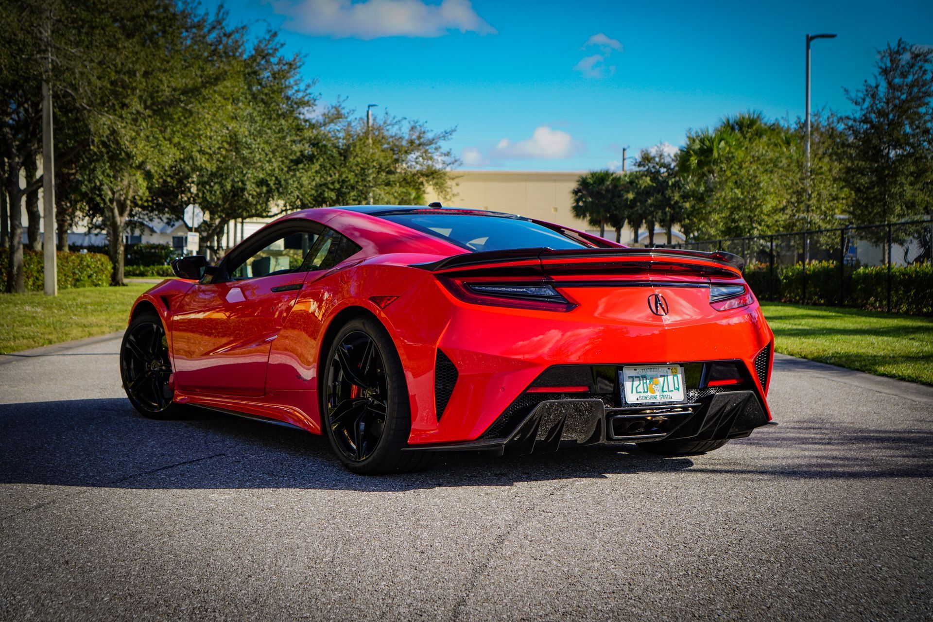 Red sports car on a road, with black wheels and a rear spoiler, in a sunny outdoor setting.