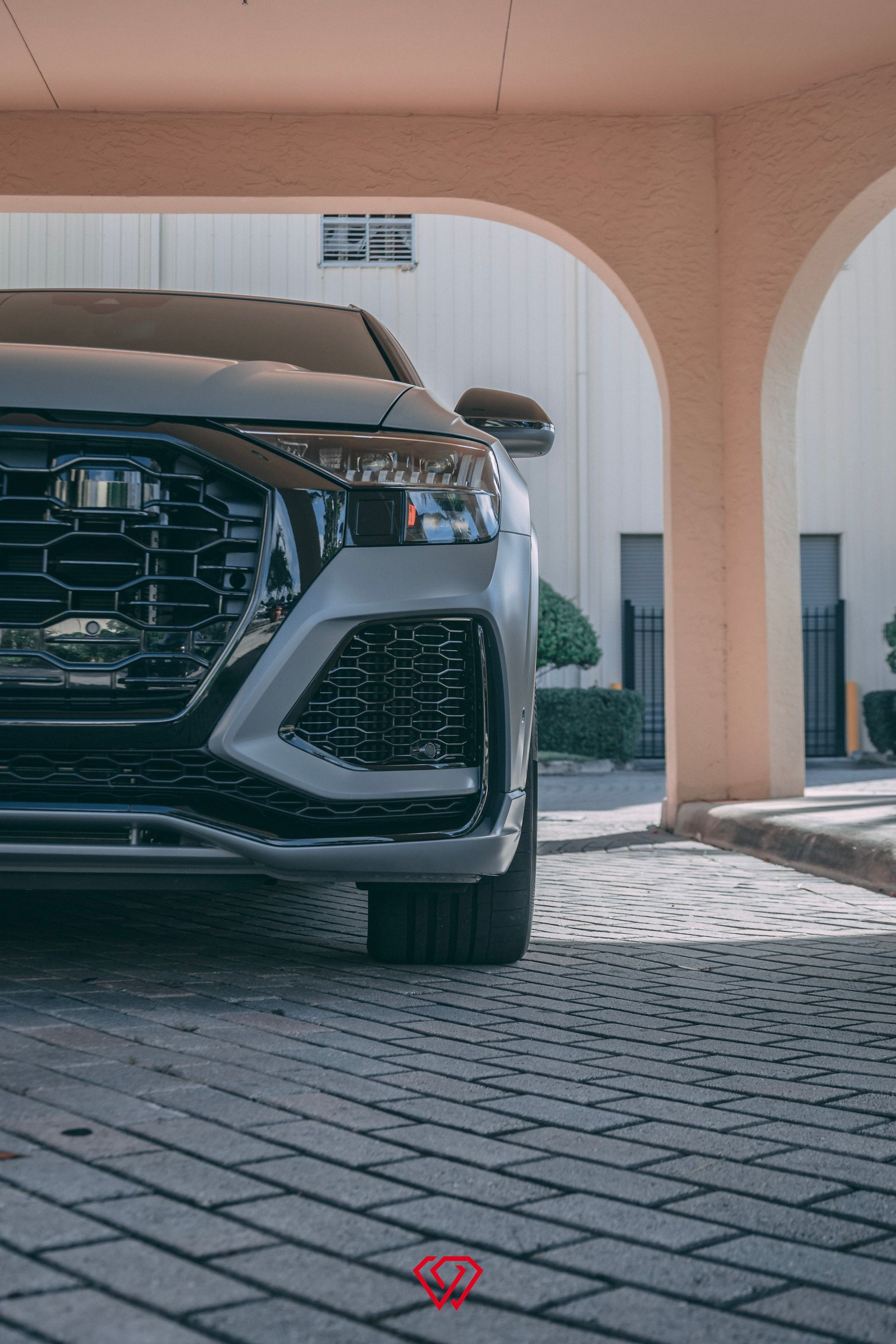 Gray Audi RS Q8 parked under an arched structure, showing the front grill and headlight. Brick pavement.