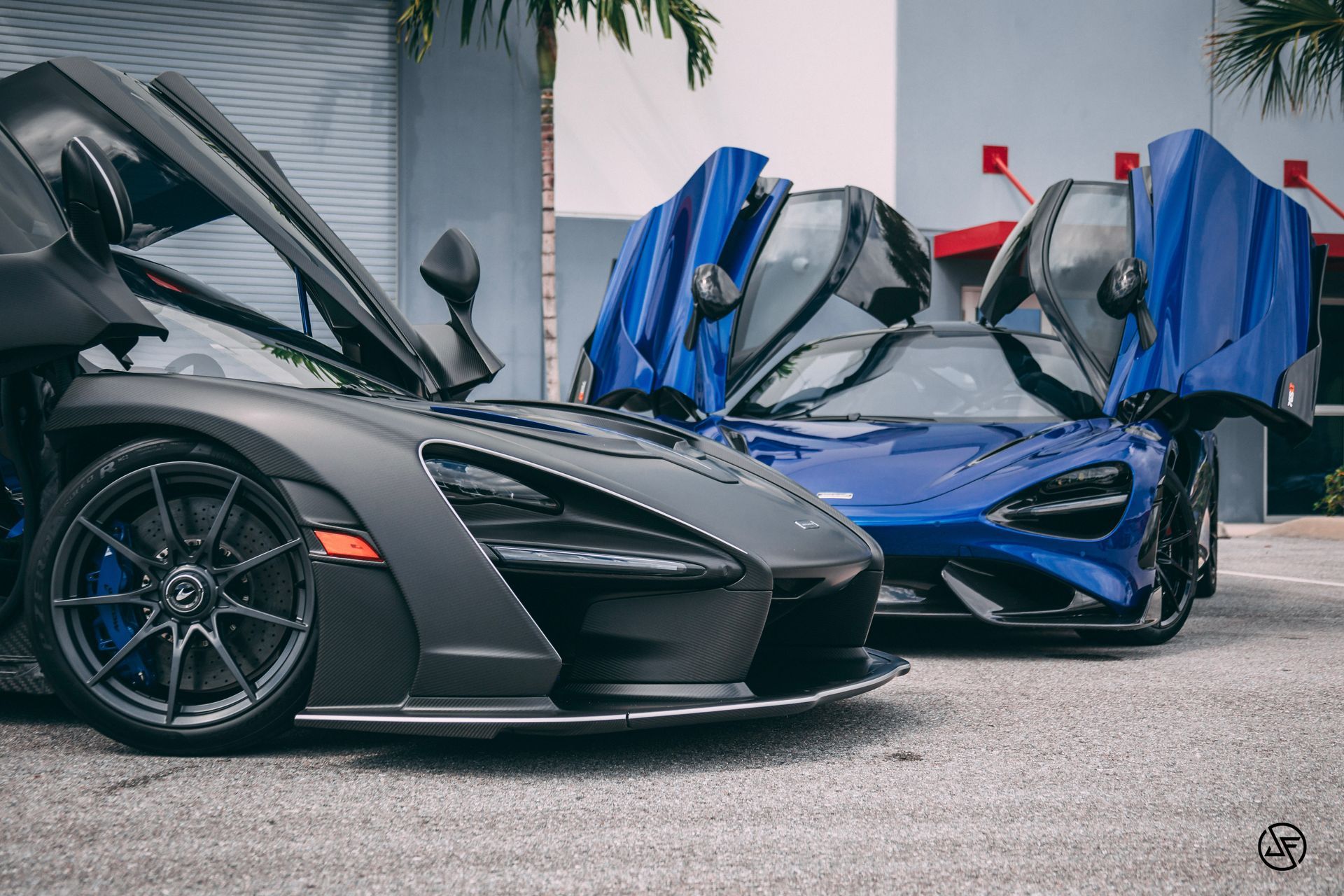 Two McLaren supercars with doors open: a gray one on the left and a blue one on the right.