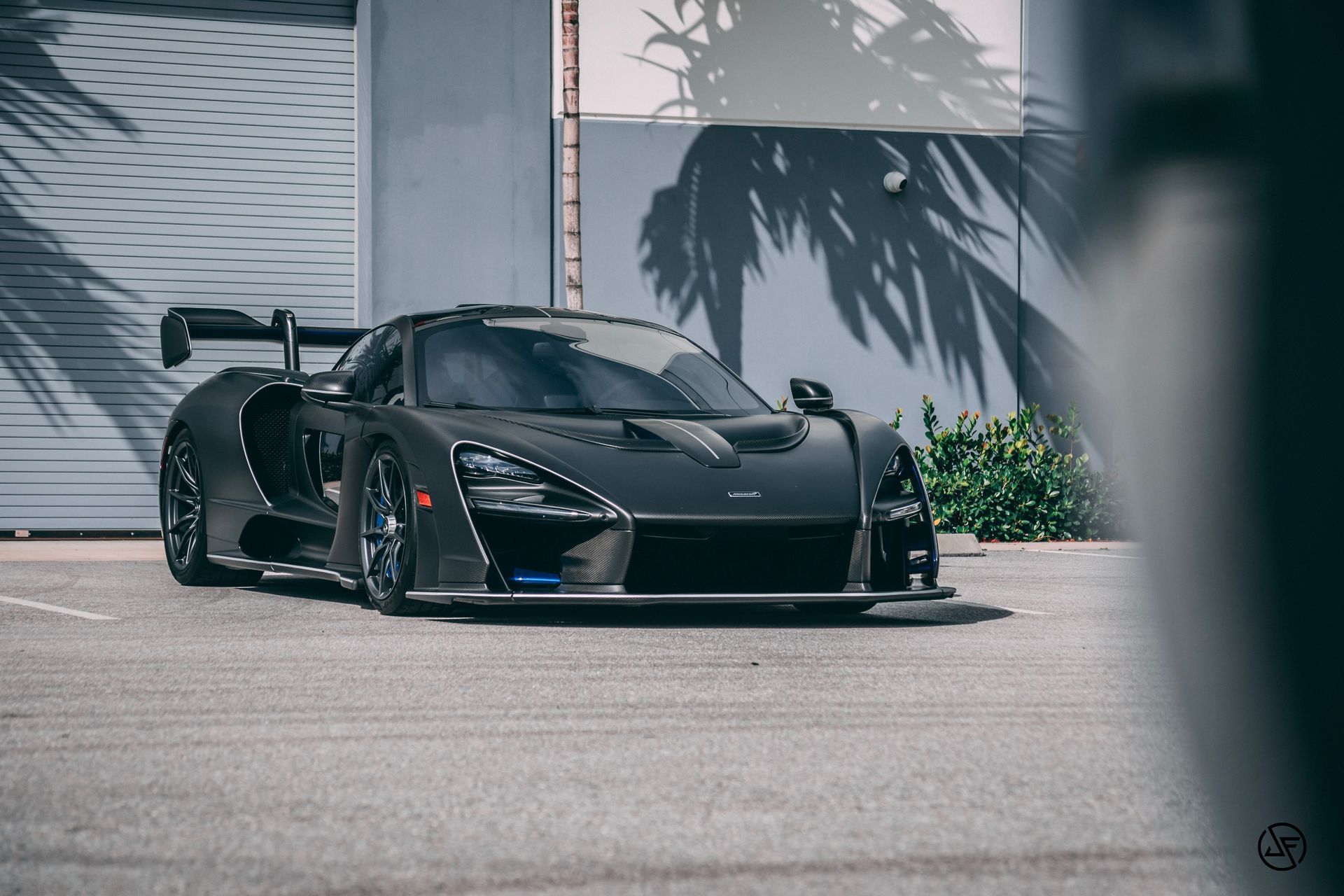 Black McLaren Senna parked on asphalt in front of a building, with palm tree shadows.