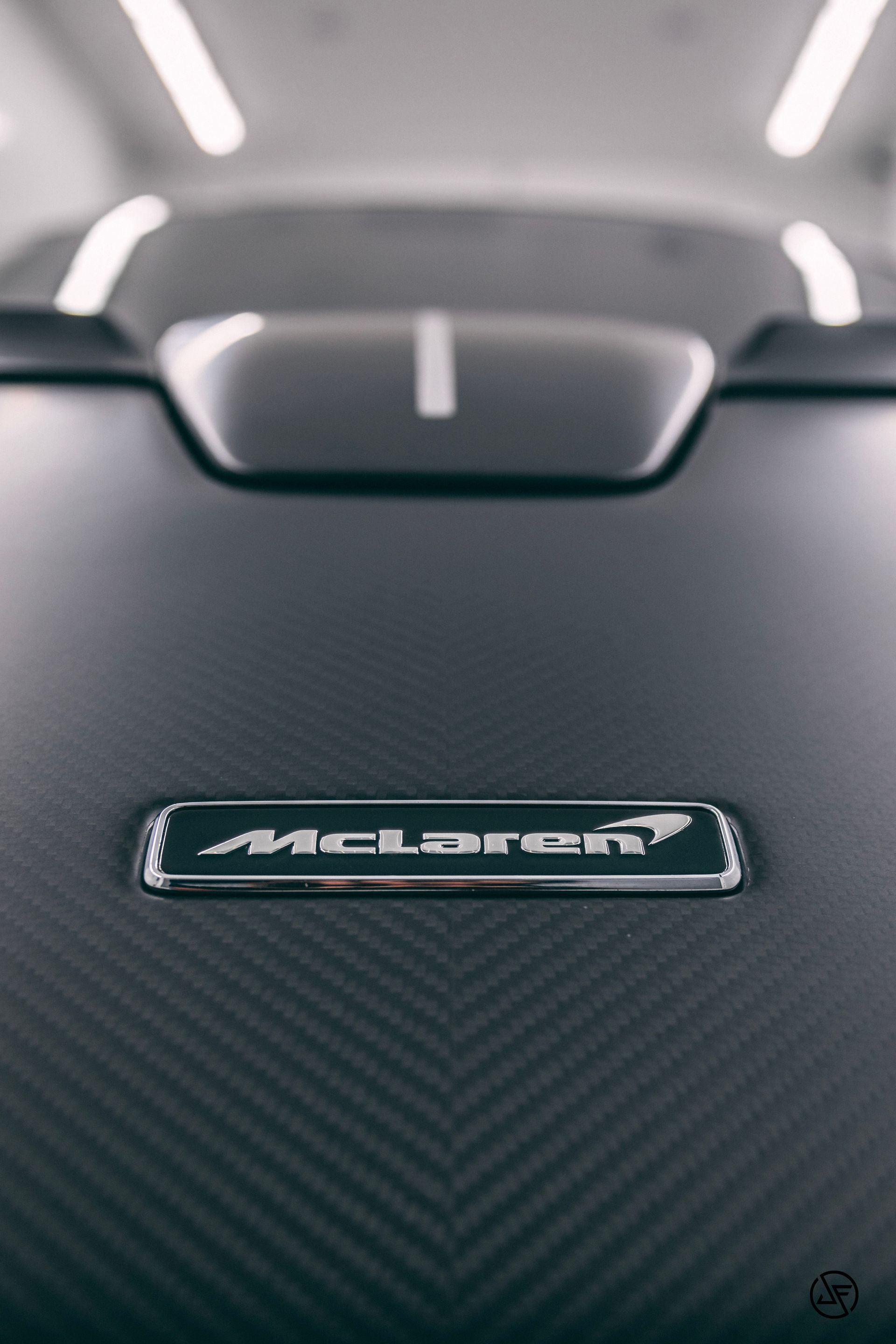 McLaren logo on a dark, carbon fiber-textured car hood, with a single white racing stripe.