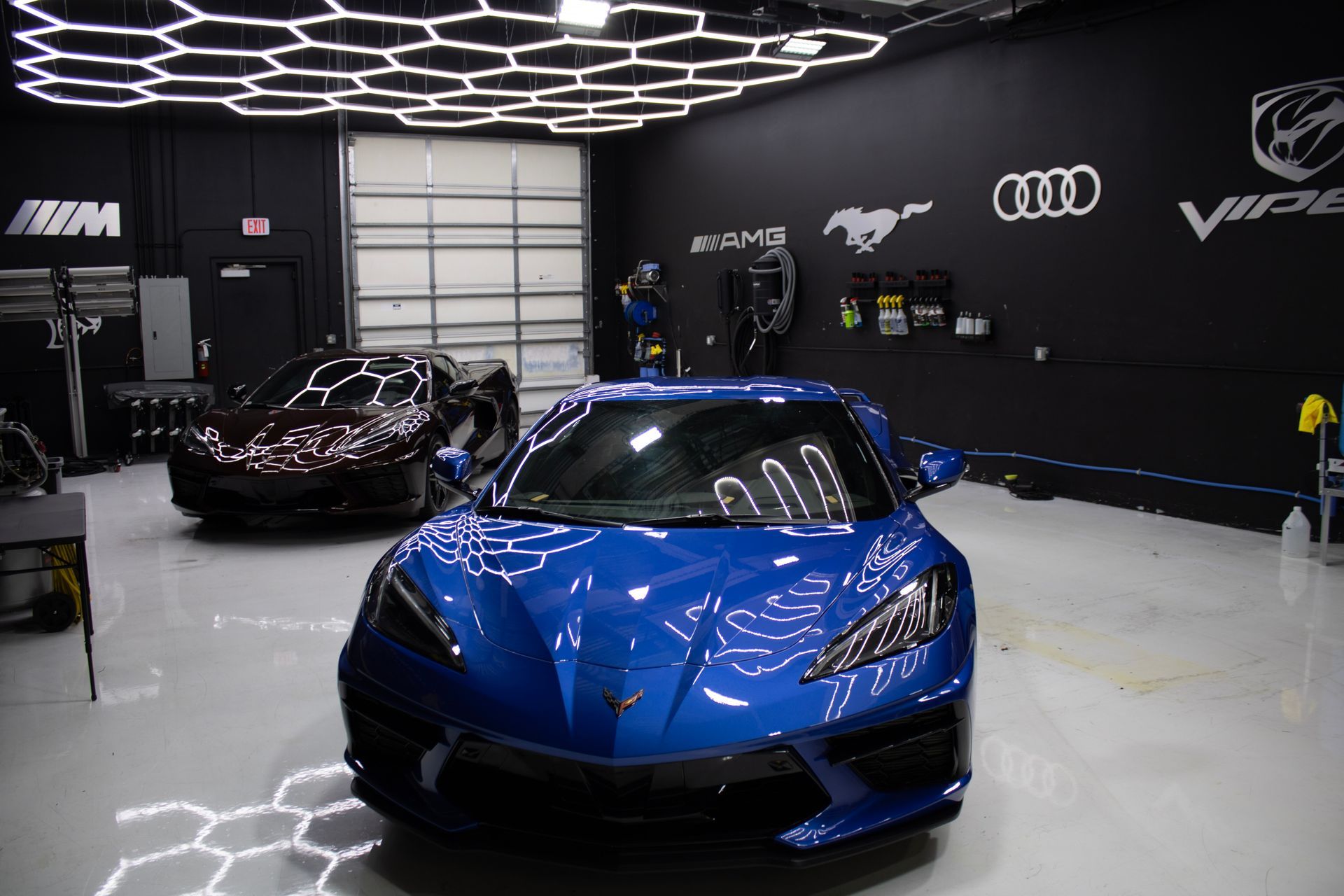 Blue sports car in a garage with honeycomb lights, black walls, and another car.