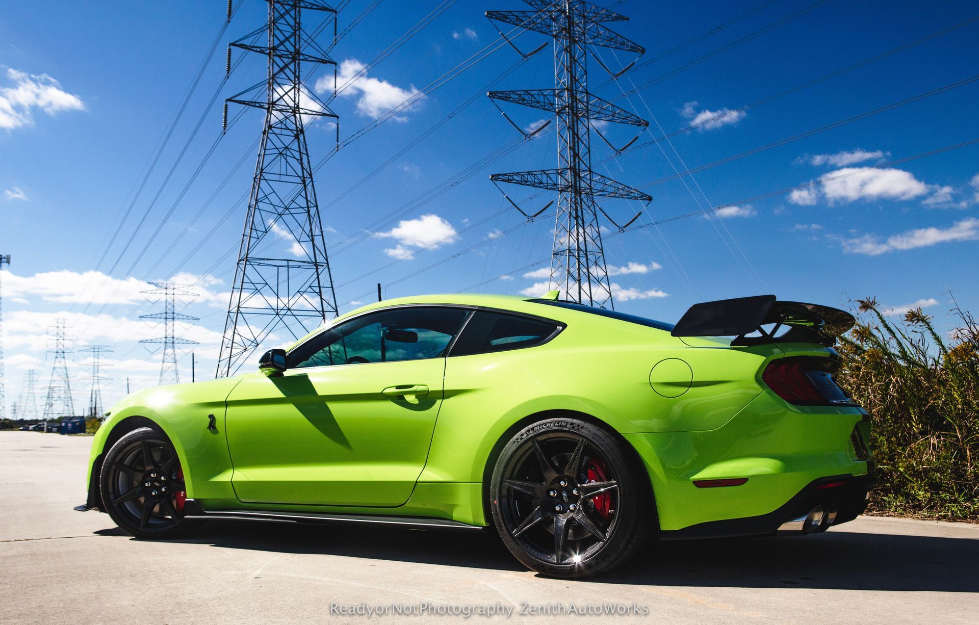 Green sports car parked, with black wheels, next to power lines under a blue sky.