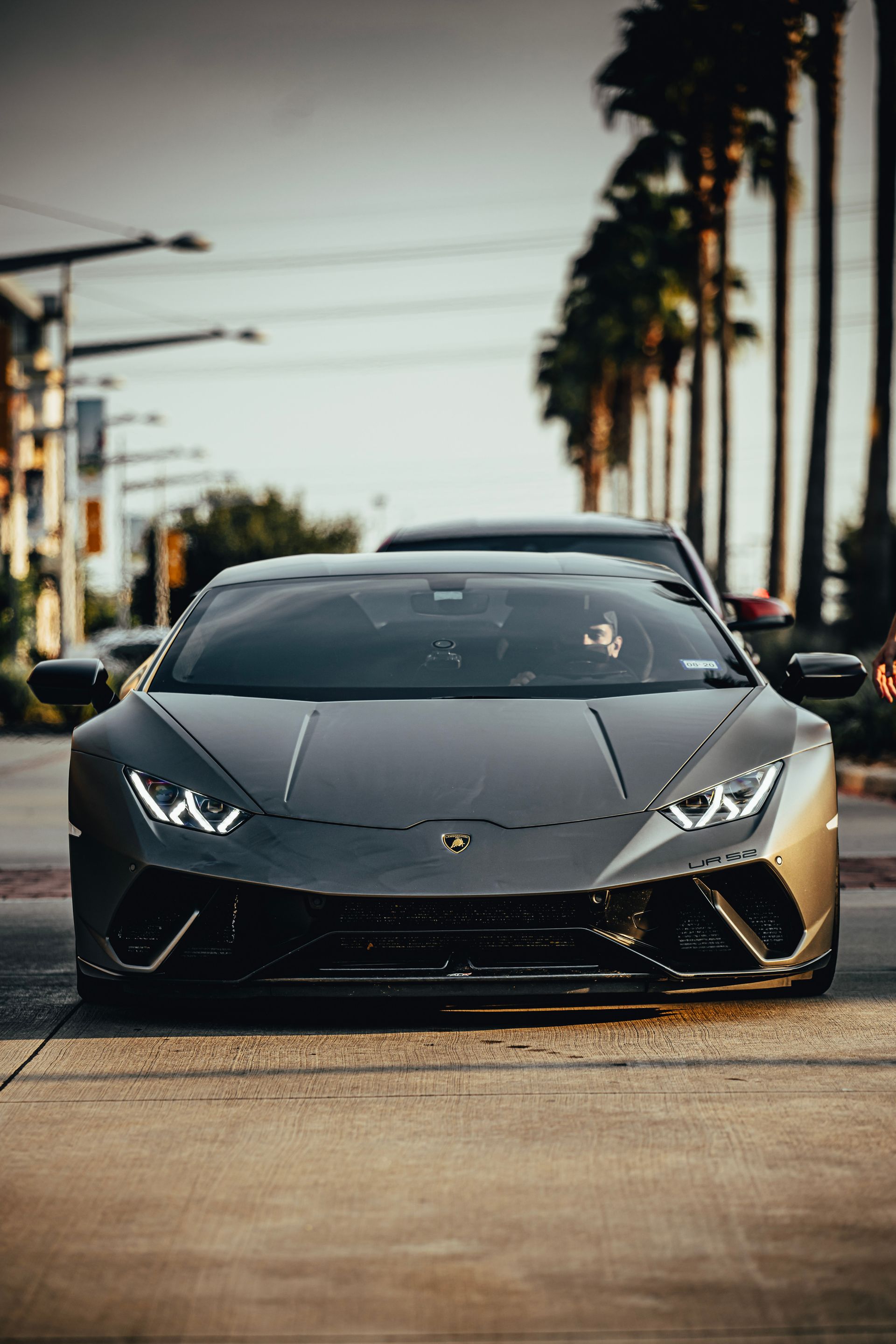 Dark gray Lamborghini Huracán on a city street, palm trees in the background.
