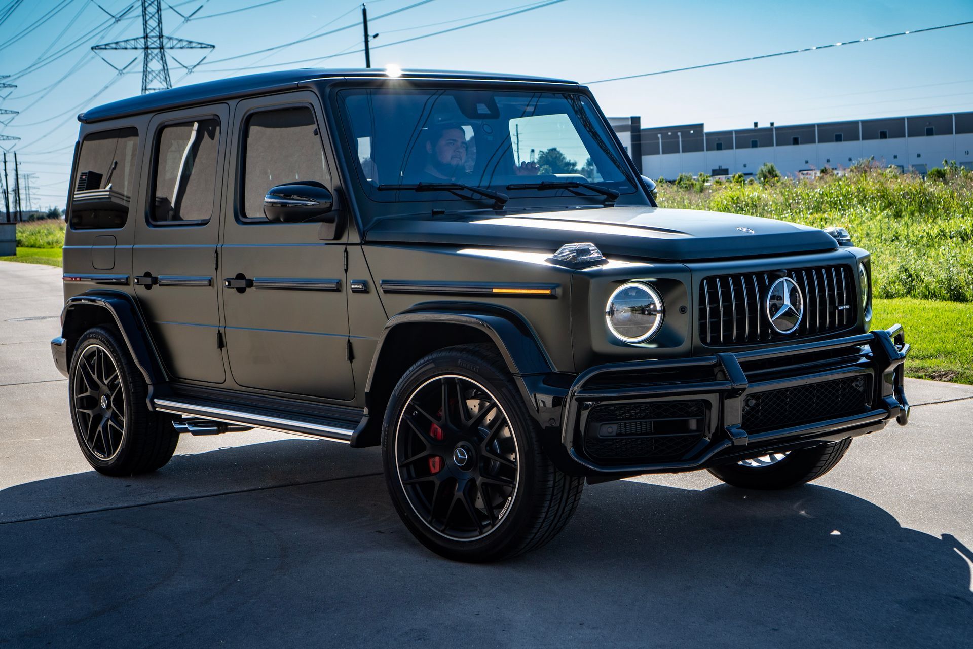 Dark gray Mercedes-Benz G-Wagon SUV parked on asphalt, near a grassy area and building, sunny day.