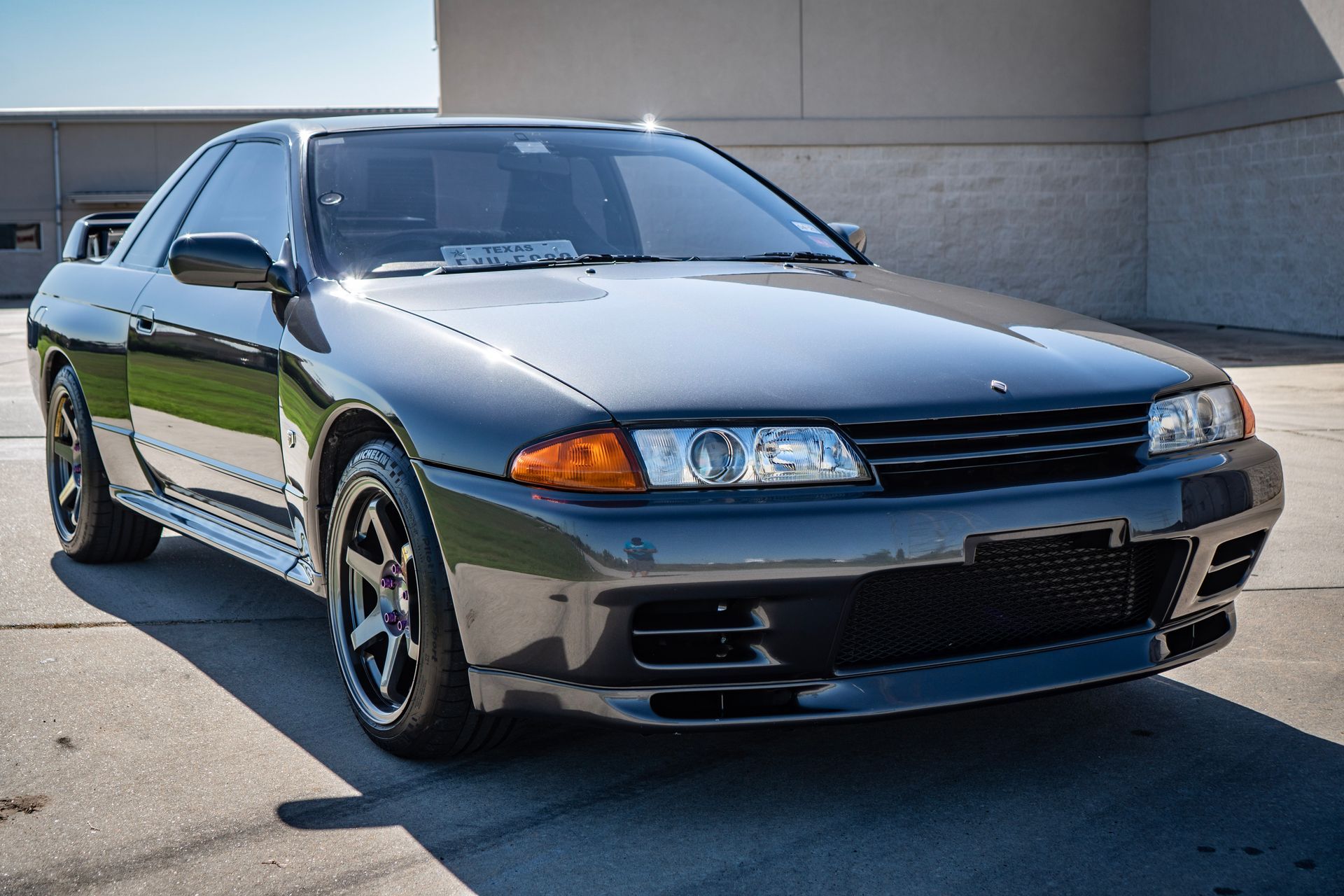 Dark gray Nissan Skyline R32 coupe parked on a paved surface, front view, sunny day.